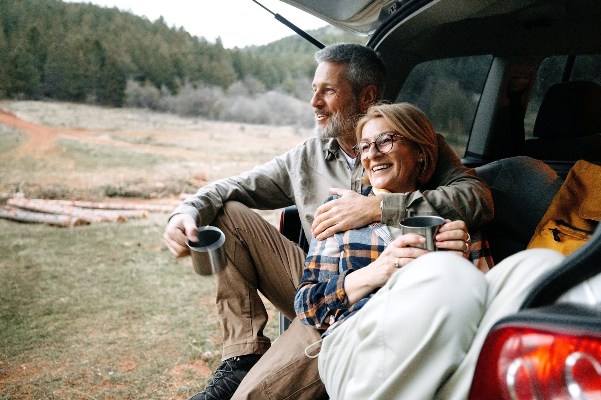 A couple sitting at the back of an SUV with mugs, smiling outdoors in the Mountain West.