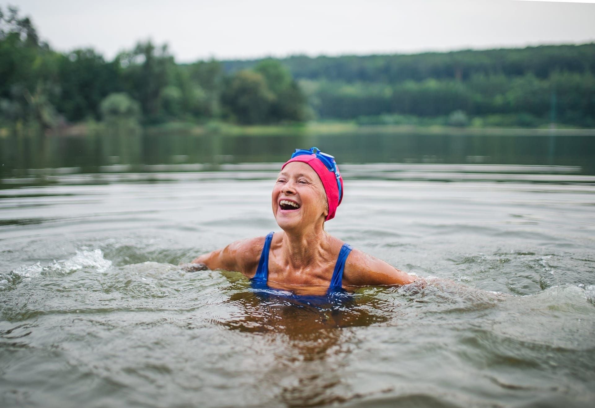 A woman laughing while swimming in a calm lake at the foot of a forested ridge.